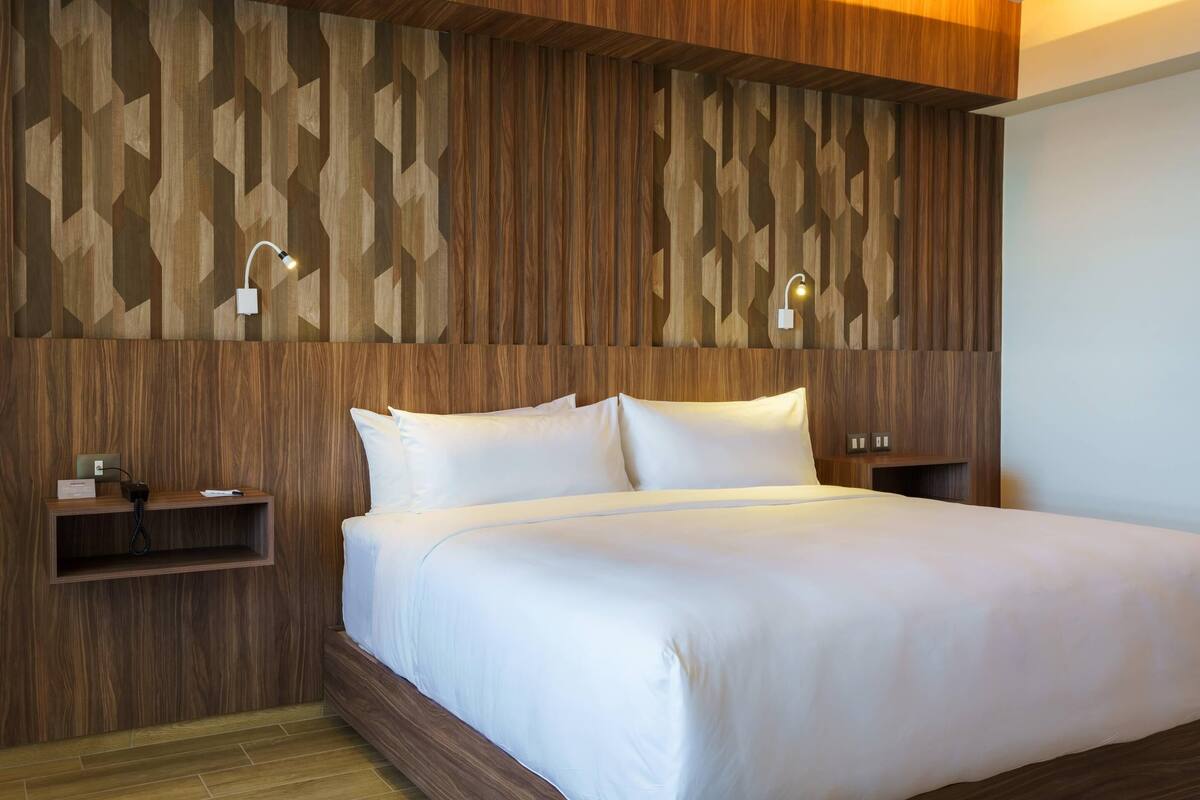 A hotel room with a wooden panel wall, a bed with white bedding, bedside tables with lamps, and a shelf with a phone and a small item.