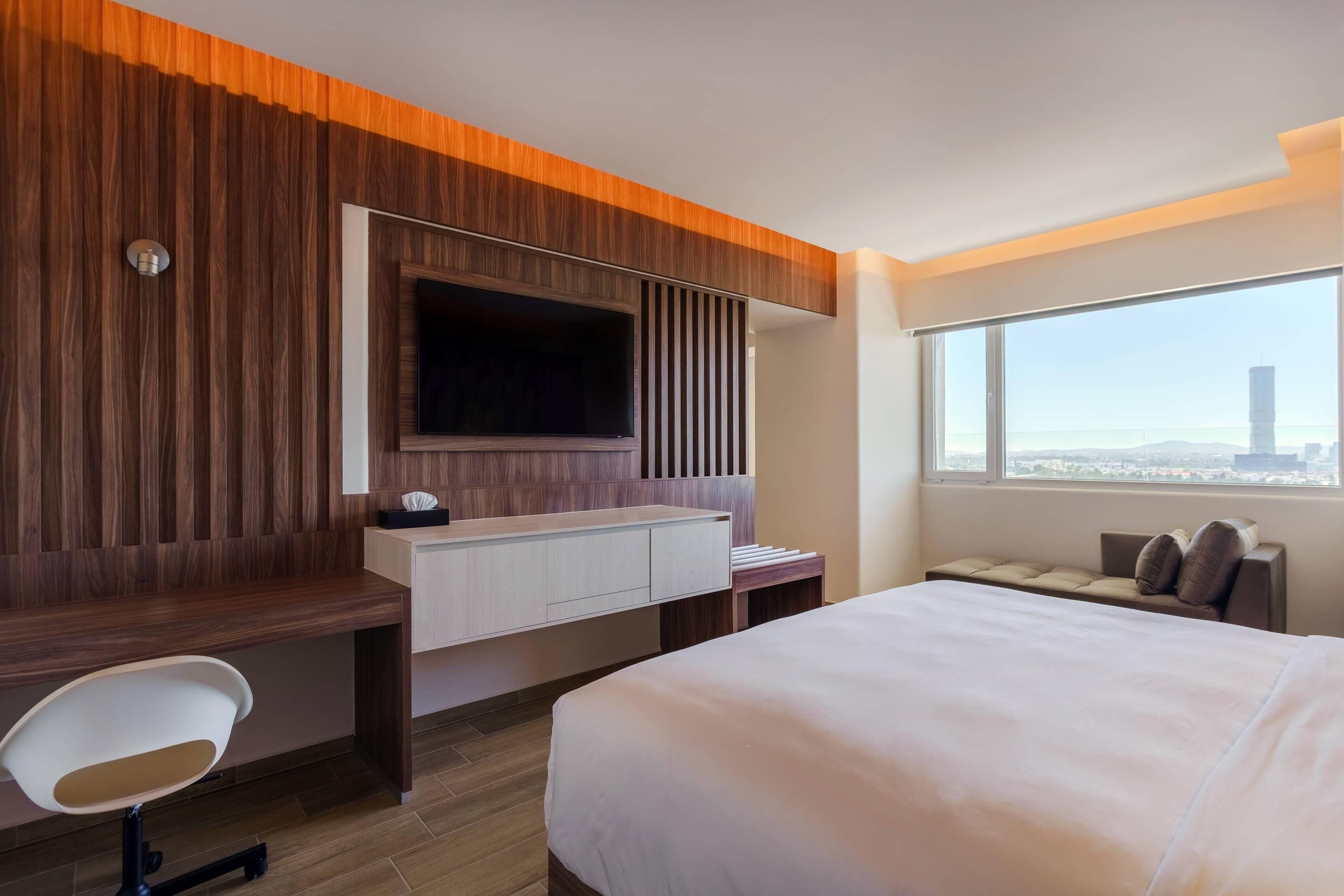 A modern hotel room with a large bed, a desk with a computer, a TV mounted on the wall, and a view of the city through a large window.