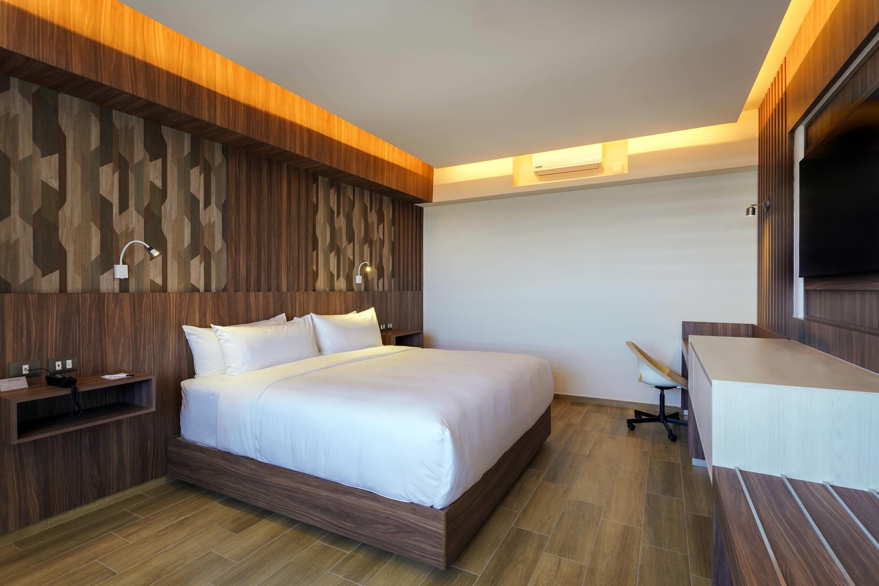 Una habitación de hotel moderna con cabecera de madera, una cama grande, un escritorio y una televisión integrada.