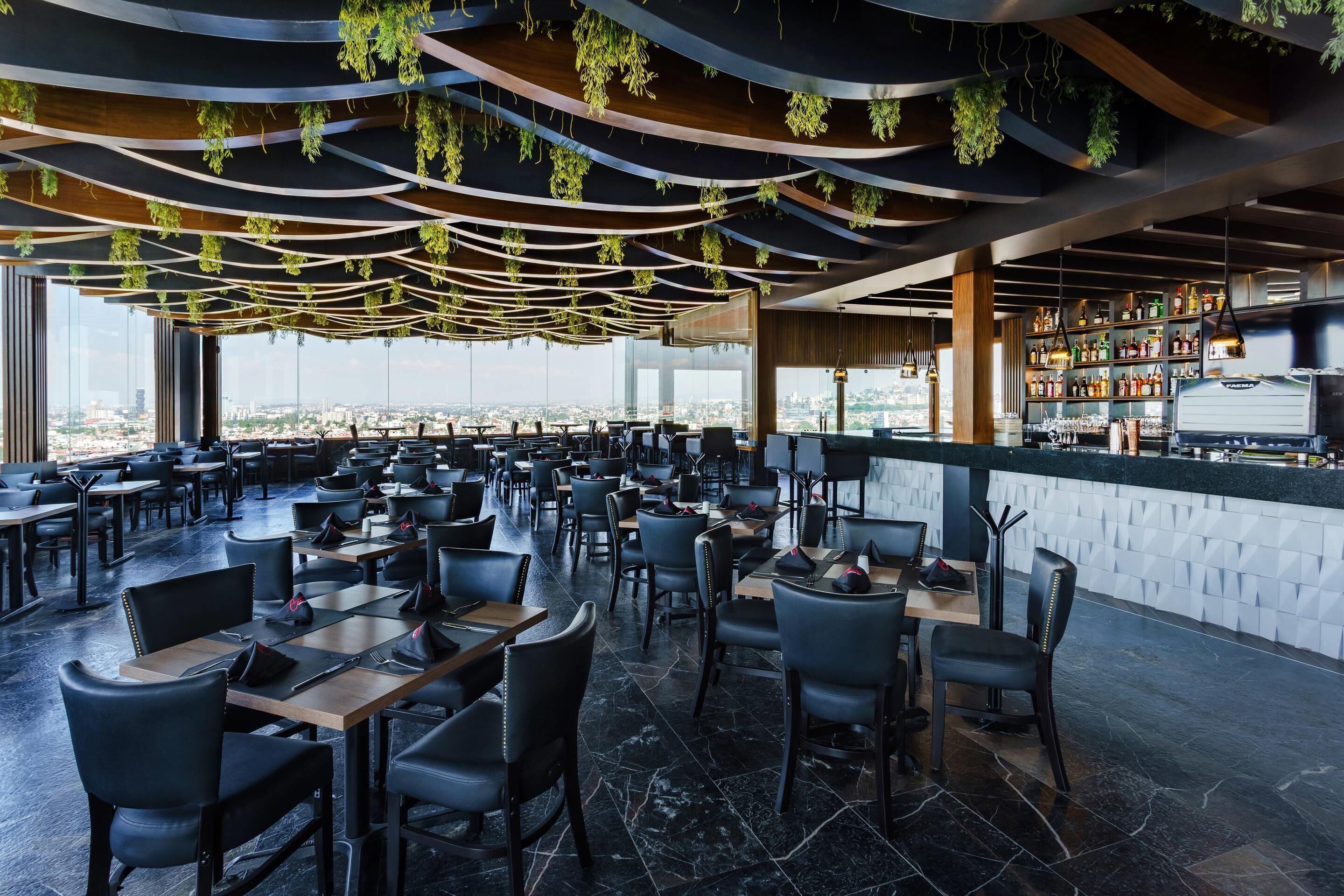 Un restaurante con techos altos, plantas colgantes y vistas a la ciudad. Las mesas y sillas están dispuestas para comer.