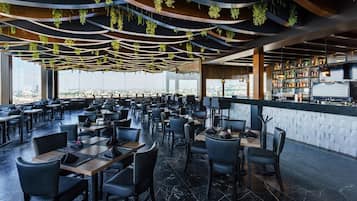 Un restaurante con techos altos, plantas colgantes y vistas a la ciudad. Las mesas y sillas están dispuestas para comer.