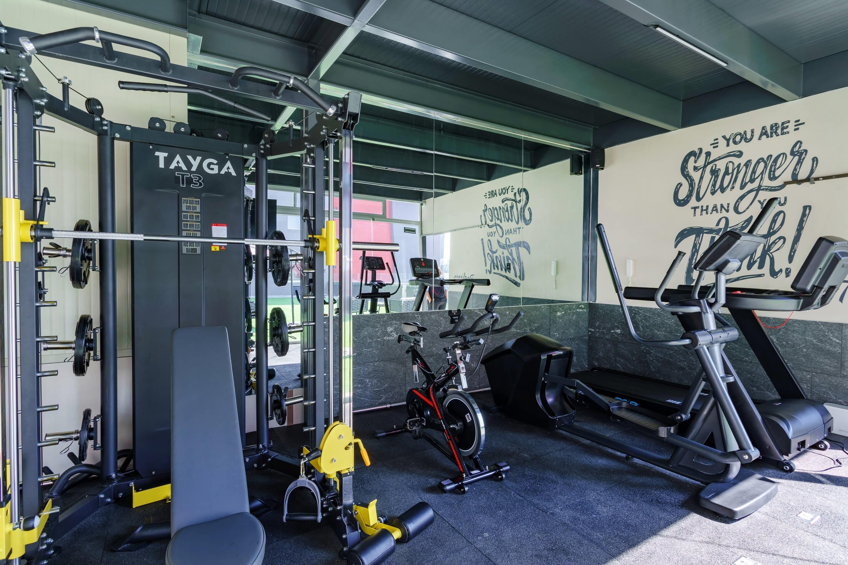 Un gimnasio con una estructura de sentadillas TAYGA T3, una prensa de banca, una bicicleta estática roja y una cinta de correr.