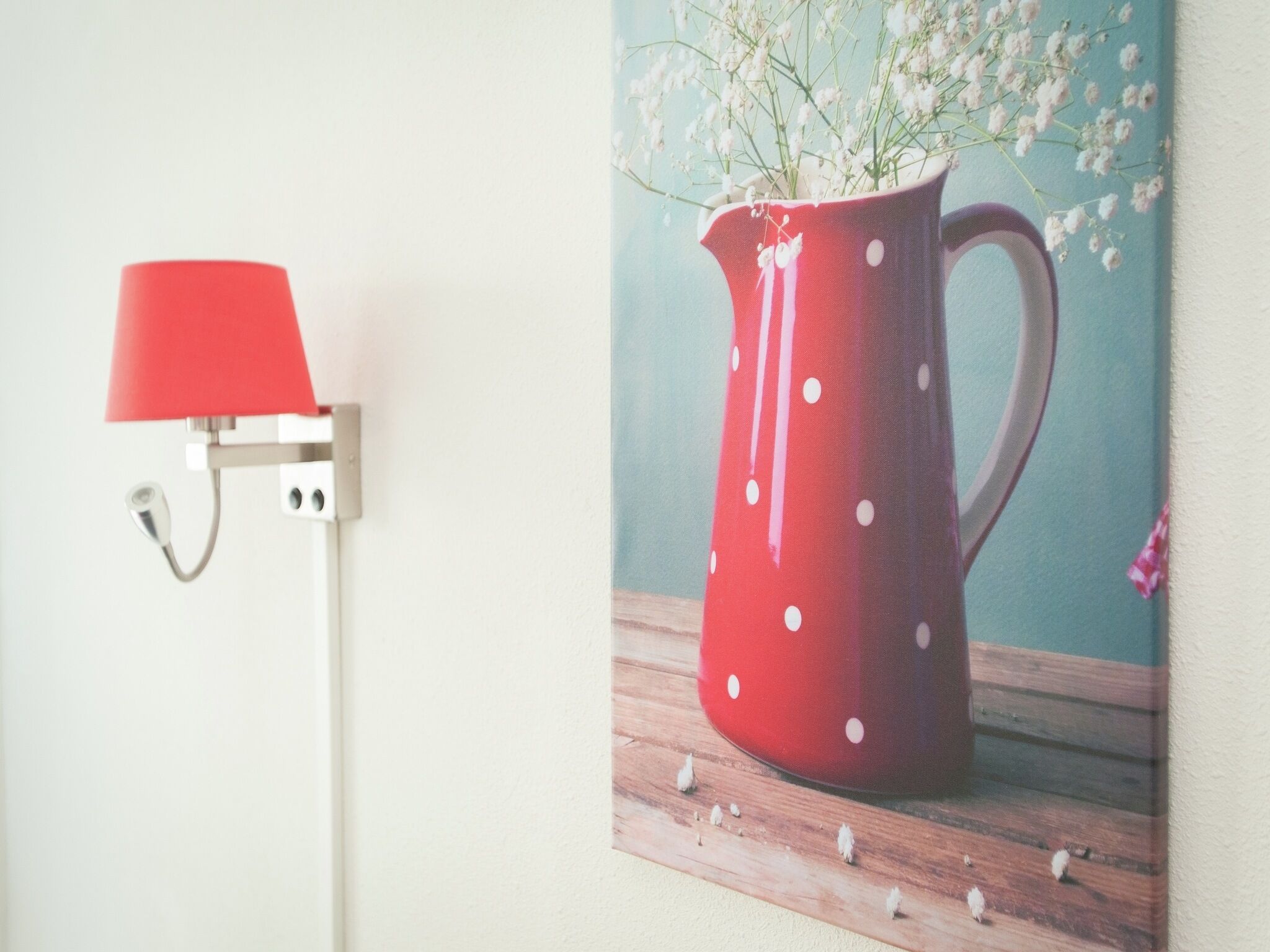 Una lámpara roja con pantalla blanca sobre una pared, una jarra roja con lunares blancos sobre una superficie de madera, y una pared azul al fondo.