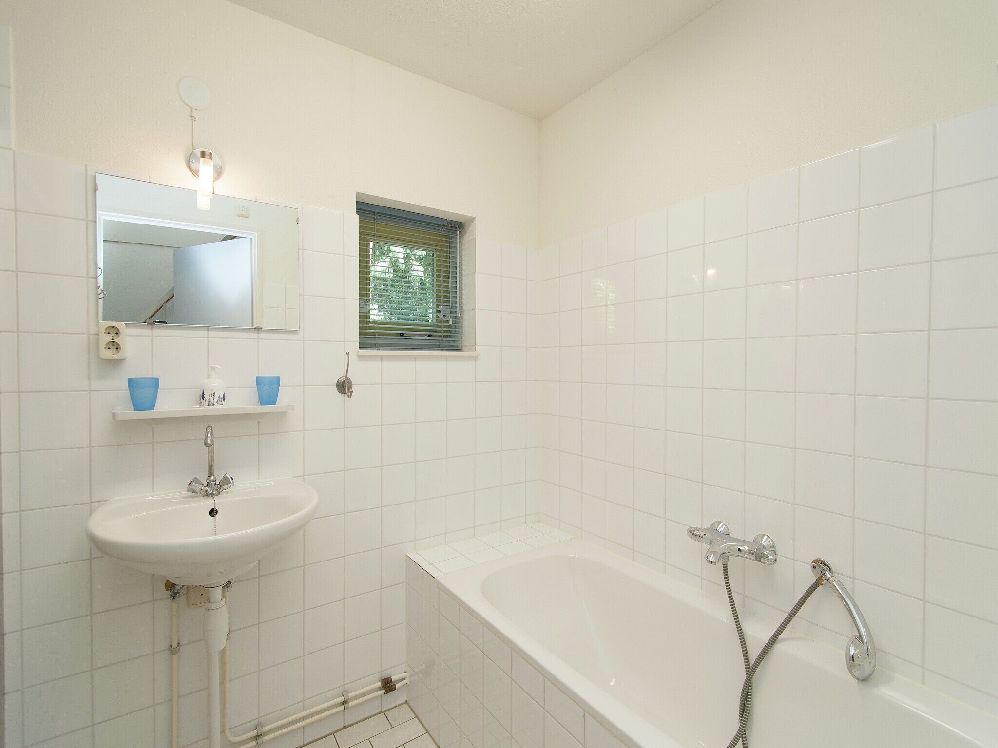 A bathroom with white tiles, a sink, a mirror, a window, and a bathtub.