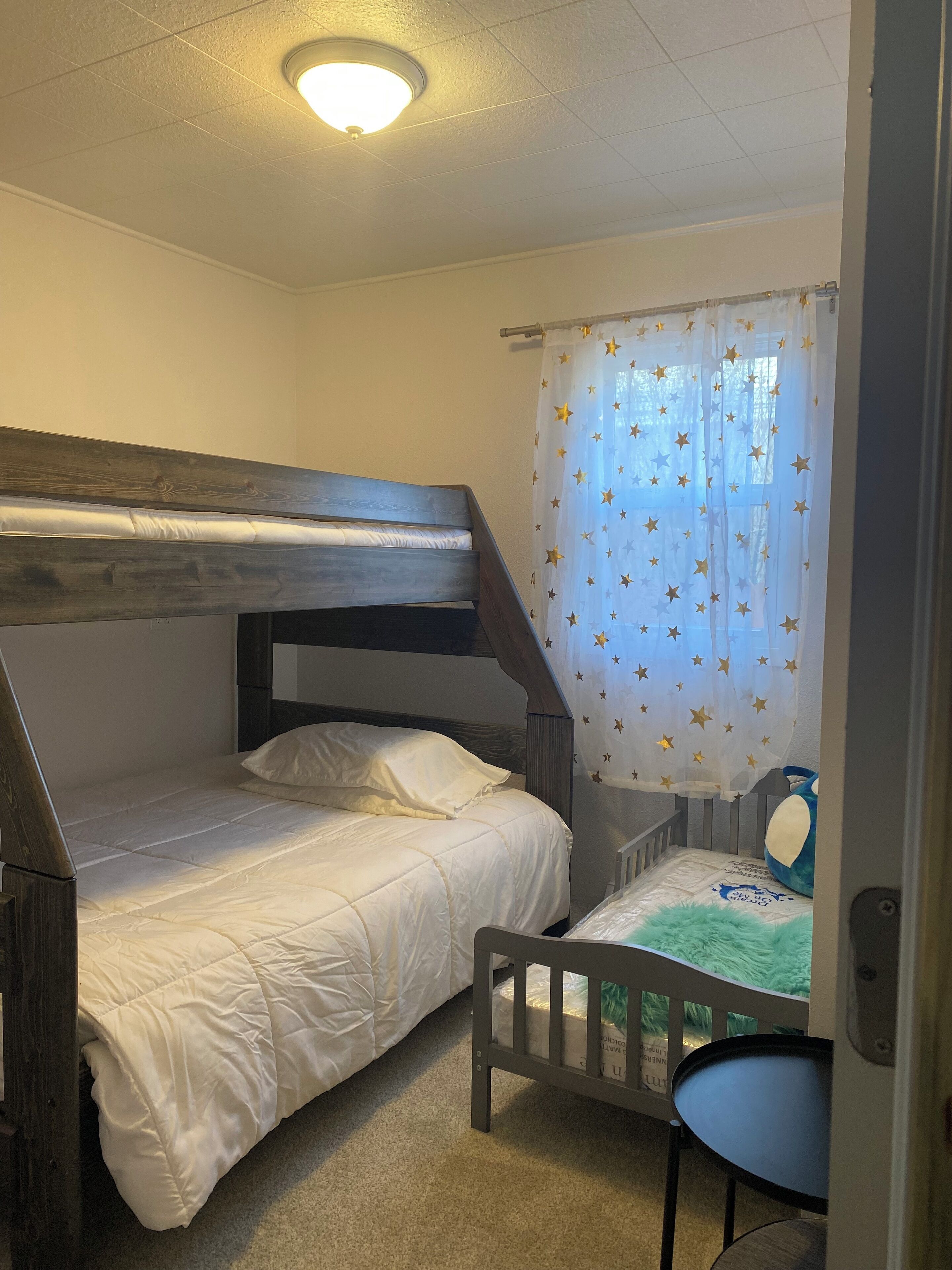 Una cama litera con cabecera de madera, un escritorio pequeño con una silla, y una ventana con cortinas estampadas de estrellas.