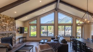 A spacious living area with a stone fireplace, large windows, a dining table, and a cozy seating arrangement. - Ski In to your Private Hot Tub. Foosball Table. (Fernie)