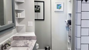 Un baño con piso a cuadros blanco y negro, un lavamanos blanco con encimera de mármol, un espejo y un ventilador de techo.