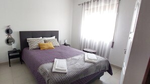 Una cama bien hecha con una colcha morada y almohadas, una mesita de noche con lámpara, y una ventana con cortinas.