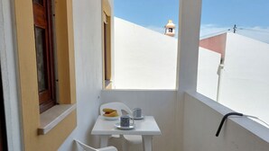 Un pequeño balcón con una mesa y silla blancos, una ventana con persianas, y vista a un edificio con tejado rojo.