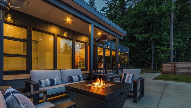 Et moderne udendørs opholdsområde med en firkantet pejsegrube, polstrede sofaer og en glasindrammet terrasse.
