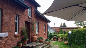 Une maison en briques avec un chemin pavé menant à l’entrée, entourée de plantes en pot et d’un grand parasol qui offre de l’ombre.