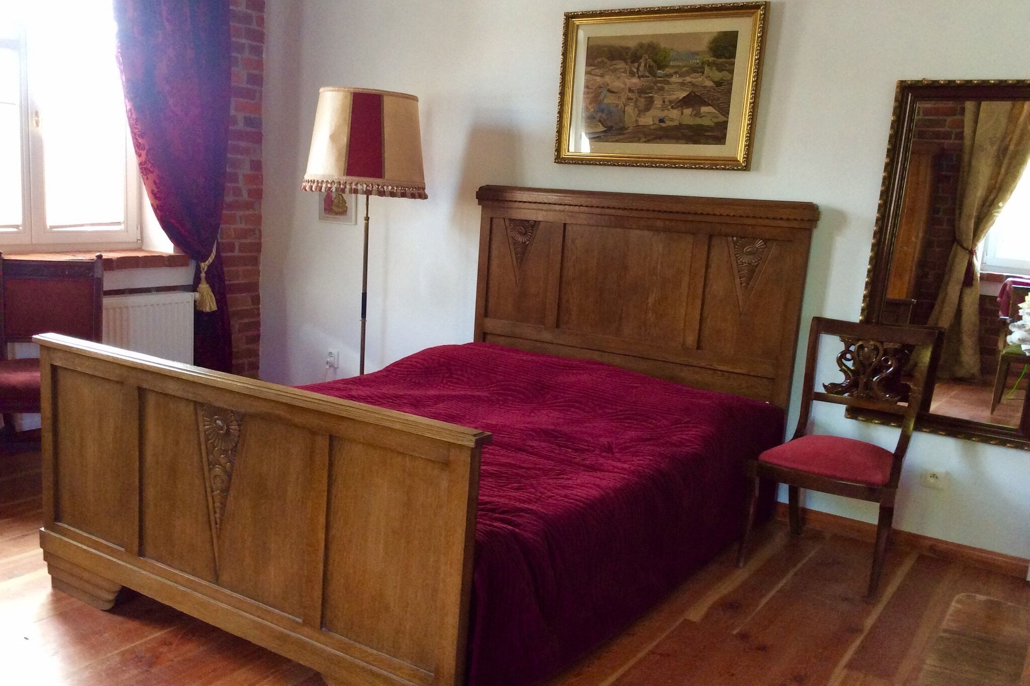 A wooden bed with a red bedspread, a wooden chair, a floor lamp, and a framed picture on the wall.