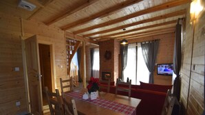 Interior de una cabaña de madera con una mesa de comedor, sillas, un sofá rojo, una chimenea y un televisor montado en la pared.
