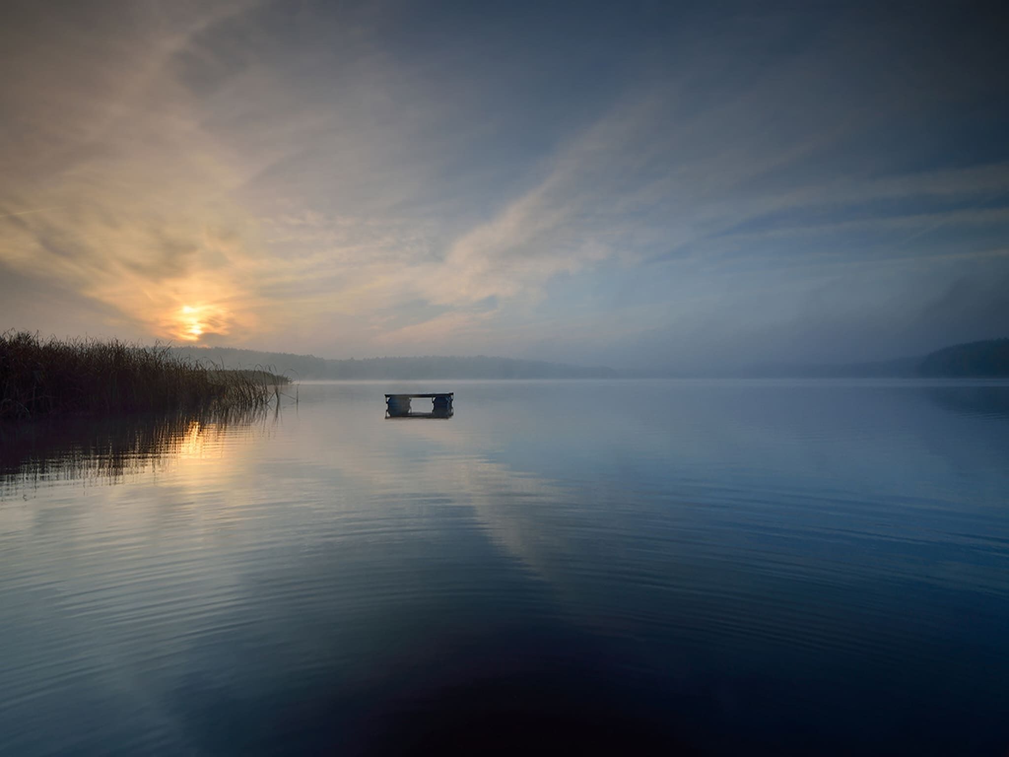 Un lac paisible, avec une jetée qui s’avance sur l’eau, entouré de roseaux et bordé d’un horizon brumeux.