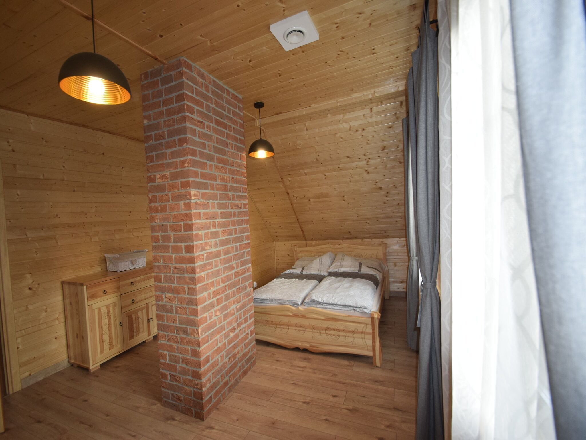 A cozy attic bedroom with a wooden ceiling, a brick chimney, a wooden bed, a nightstand, and a hanging light fixture.
