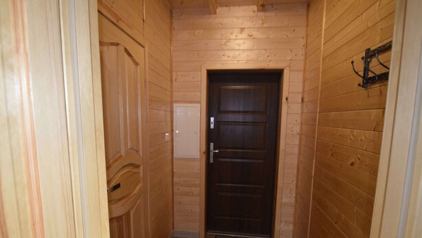 Un estrecho pasillo de madera con una puerta de madera oscura al final, un pequeño enchufe eléctrico y un gancho metálico en la pared.