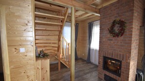 Interior de una cabaña de troncos con escalera de madera, chimenea y vista al exterior a través de una ventana.