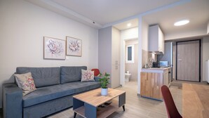 A modern living room with a grey sofa, a wooden coffee table, and a red chair. There is a kitchen area with a wooden island and cabinets. - Ilisia 2 person apartment by MPS (Athina)