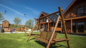 Un ensemble de balançoires en bois et un hamac sont installés dans une zone herbeuse, avec une cabane en bois et un bâtiment doté d’un balcon en arrière-plan.