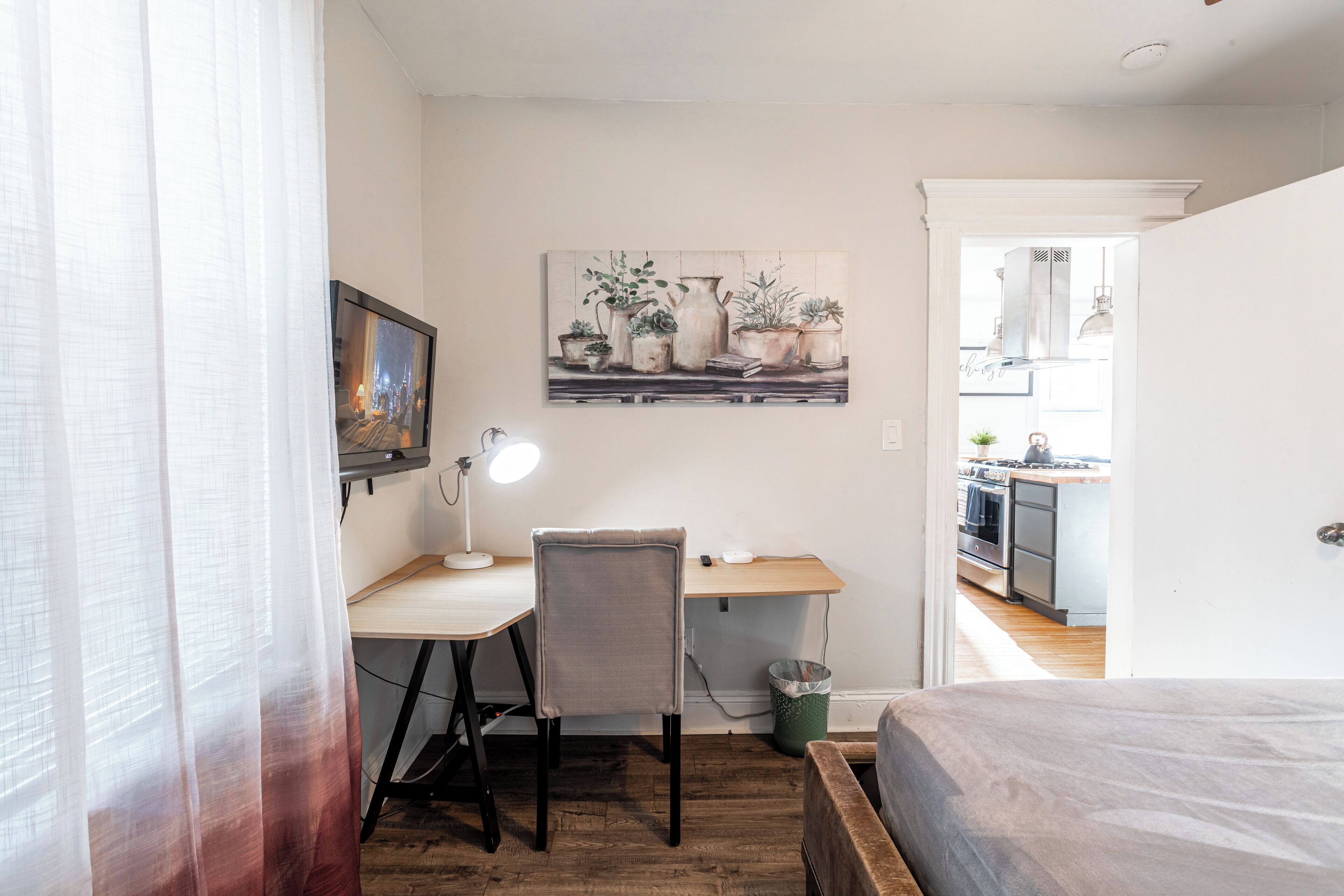 A bedroom with a wooden bed, a desk with a chair, a lamp, and a painting of plants on the wall.