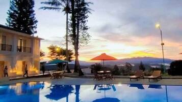 A swimming pool with lounge chairs and umbrellas, surrounded by tall trees and a sunset view.