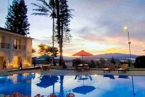 A swimming pool with lounge chairs and umbrellas, surrounded by tall trees and a sunset view. - The Balcone Suites & Resort (Gadut)