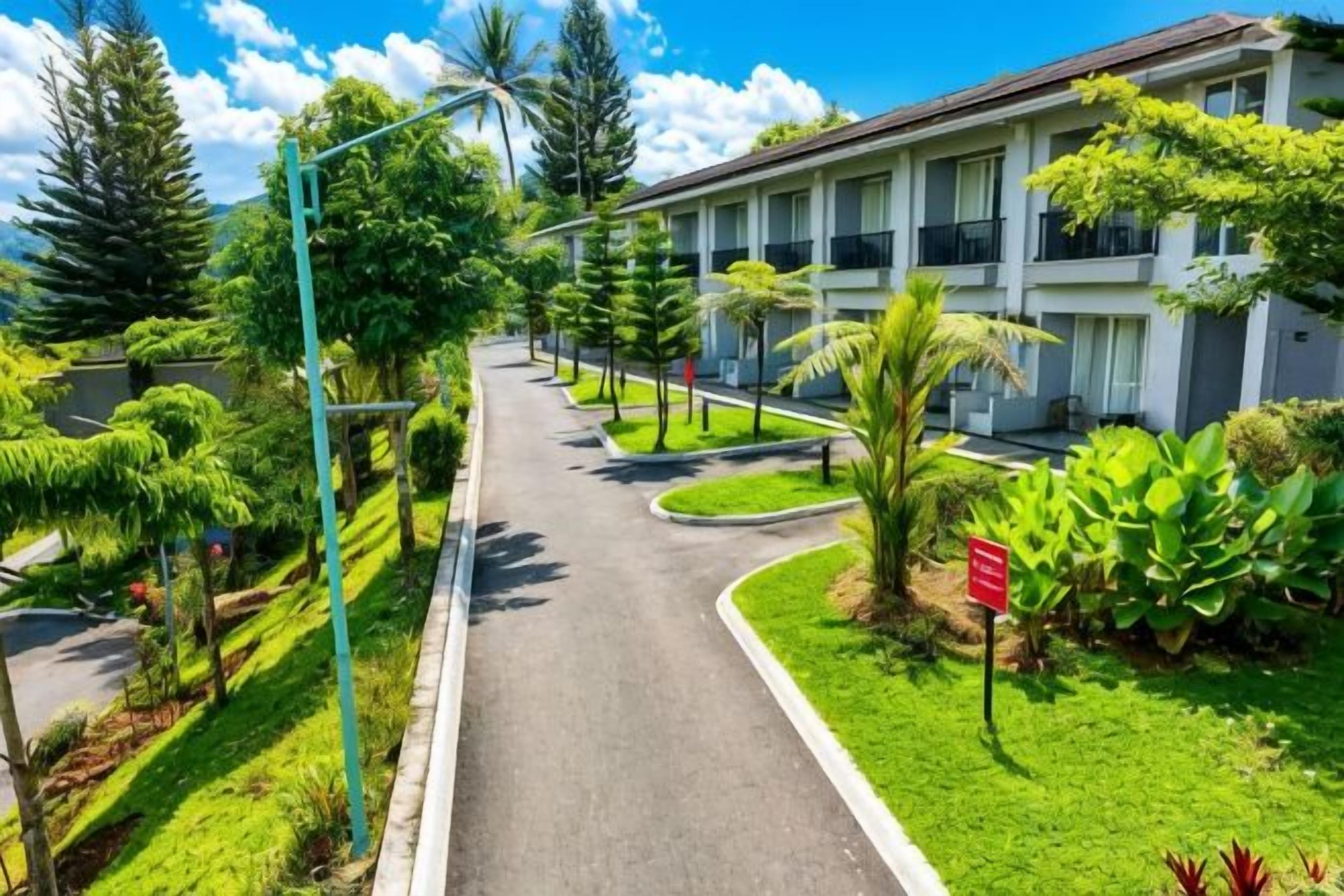 A pathway leading to a building with multiple balconies, surrounded by lush greenery and a clear sky.