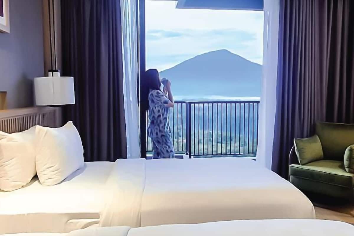 Une chambre d’hôtel avec un grand lit, un fauteuil vert et un balcon offrant une vue sur la montagne.