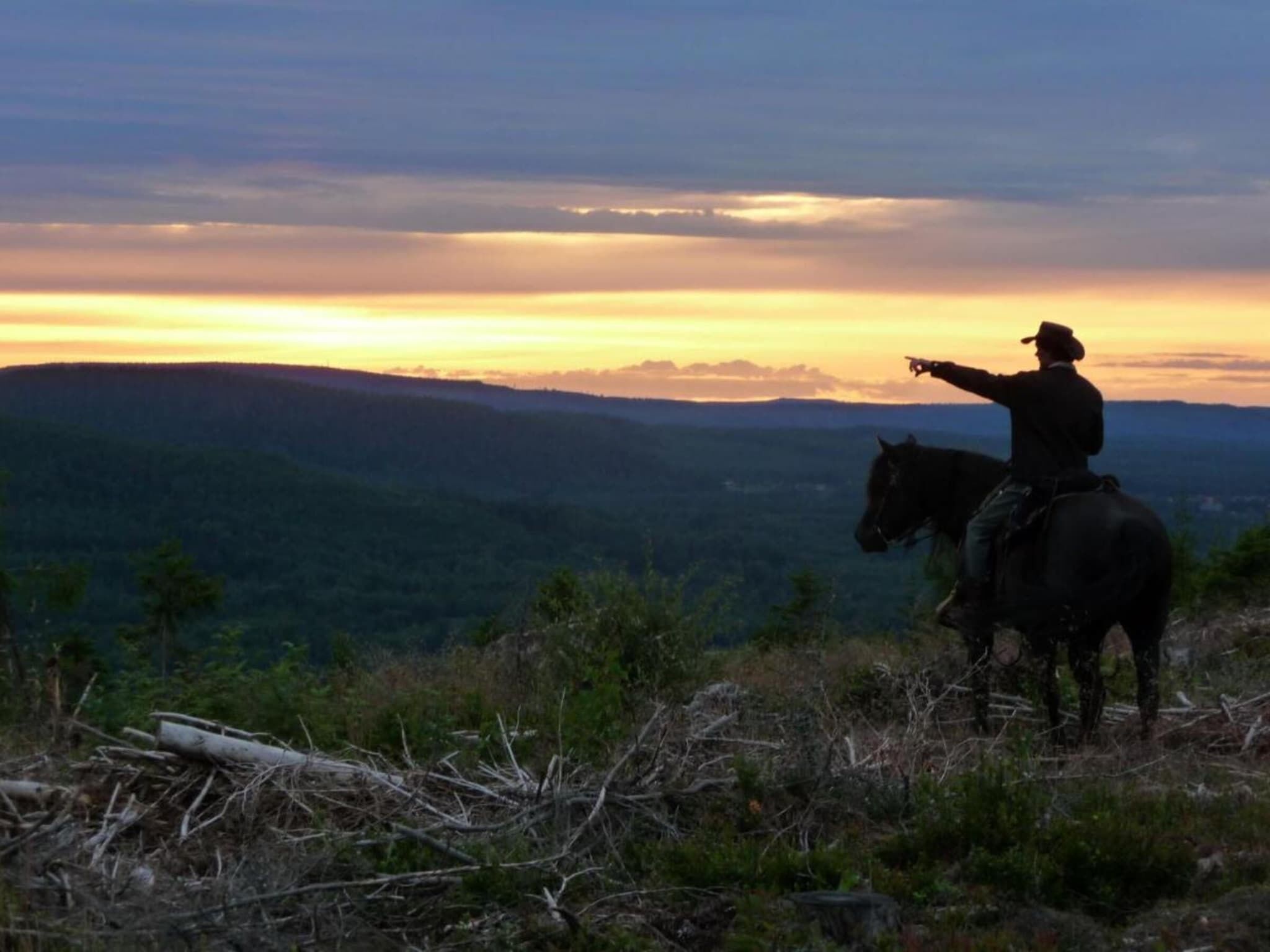 Una persona a caballo señalando hacia un atardecer sobre un paisaje montañoso.
