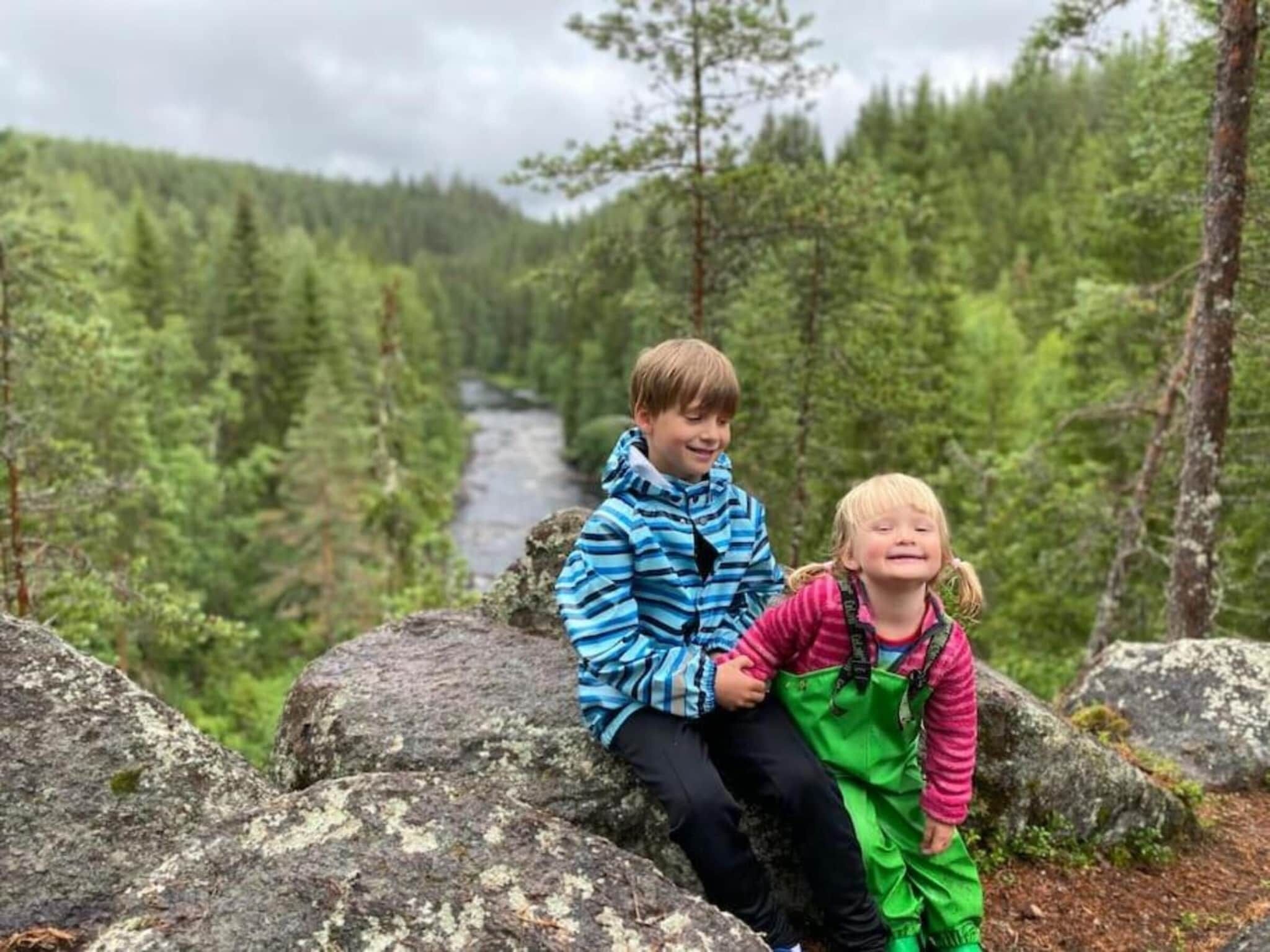 Two children sitting on a rock with a forest and a river in the background.