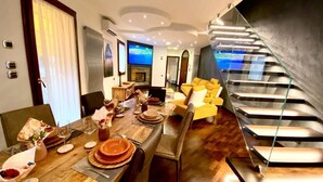 Un comedor moderno con una mesa de madera dispuesta para una comida, un sofá con cojines amarillos y una escalera con barandas de vidrio.