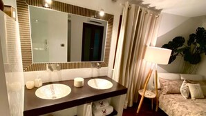 Un baño con dos lavamanos, un espejo grande y una ducha. Hay una cama con una lámpara y una planta en la esquina.