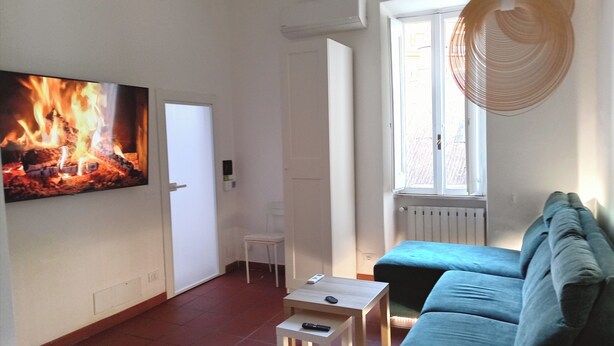 A living room with a fireplace, a sofa, a small table, and a television displaying a fire. - Your Home In Trastevere (Roma)