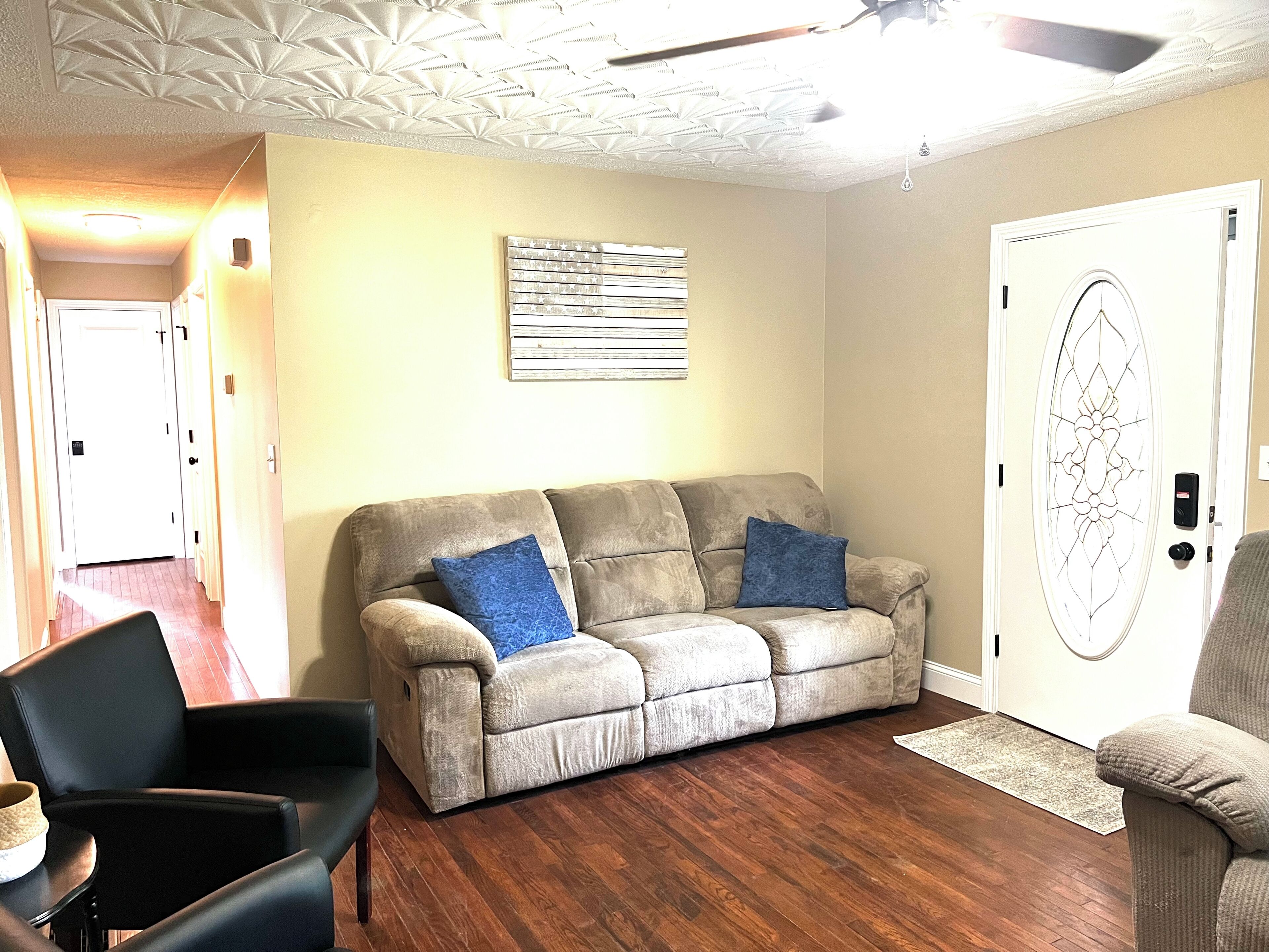 Una sala de estar con un sofá color beige, dos cojines azules, un sillón negro y piso de madera.