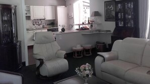 Un salon avec deux fauteuils blancs, une table basse ornée d’un vase, et une vue sur la cuisine.