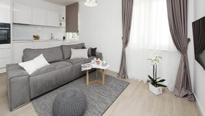 Un salon moderne avec un canapé gris, une table basse et une petite table d’appoint. On y trouve également un coin cuisine avec des meubles blancs et une fenêtre agrémentée de rideaux.