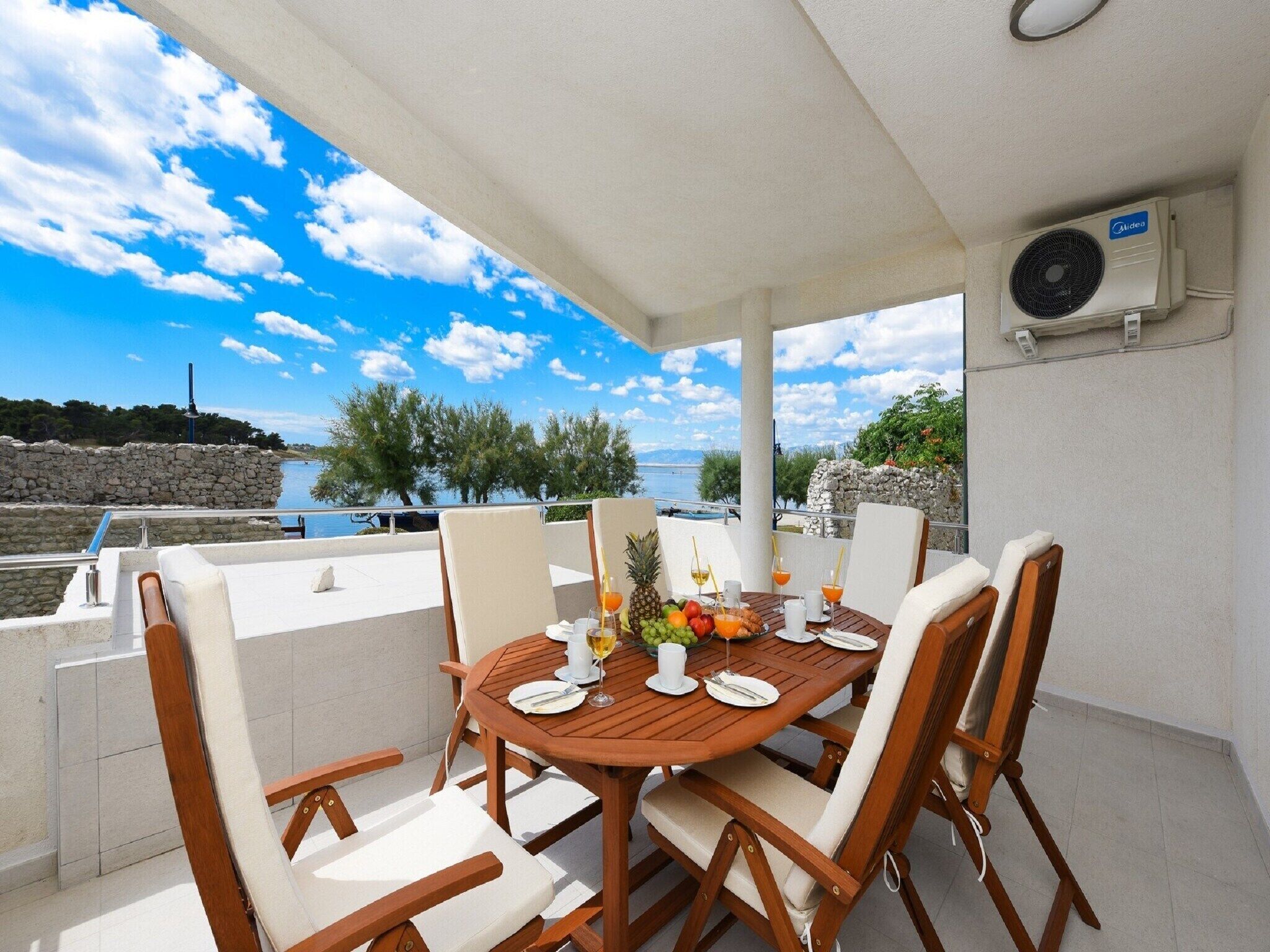 Un área de comedor al aire libre techada, con una mesa lista para cuatro personas, rodeada de sillas y vista a una masa de agua con un muro de piedra y vegetación.