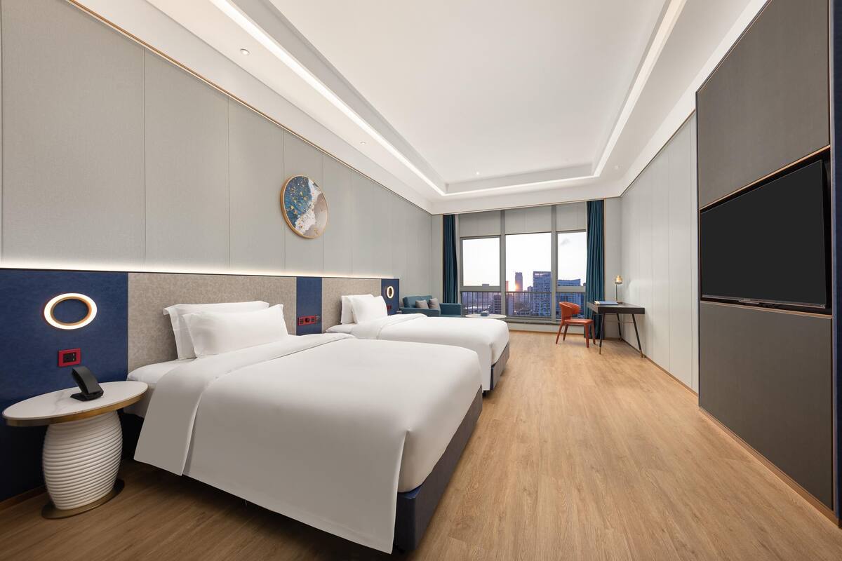 A hotel room with two beds, a desk, a chair, a TV, and a view of the city.