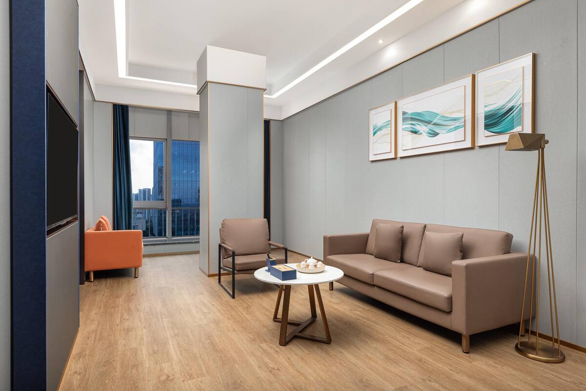 A modern hotel room with a sofa, armchair, and coffee table. There is a large window with a view of a cityscape.