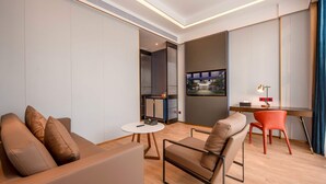A modern hotel room with a sofa, a small round table, a desk with a chair, and a view of the city through a large window. - Ramada BY Wyndham Guangzhou Huangpu (Guangzhou)