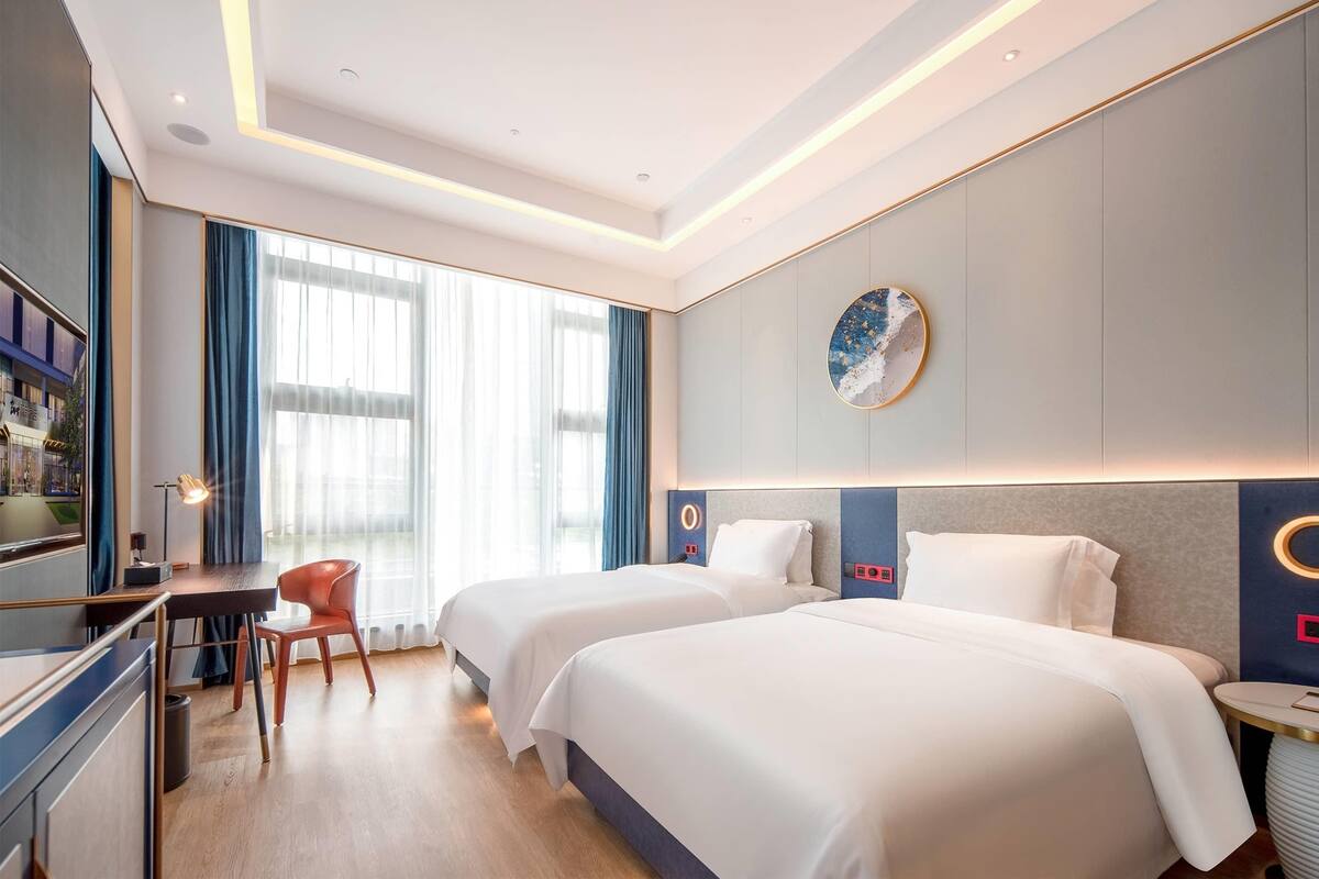 A hotel room with two beds, a desk, a chair, a TV, and a large window with curtains.