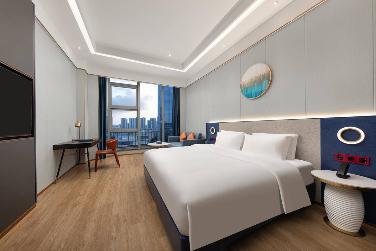 A modern hotel room with a large bed, a desk, a chair, and a view of the city through large windows.