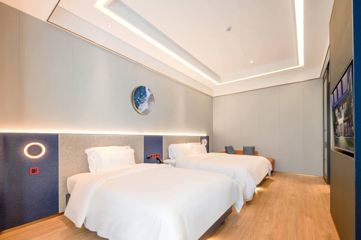 A modern hotel room with two beds, a round wall clock, and a built-in bookshelf.