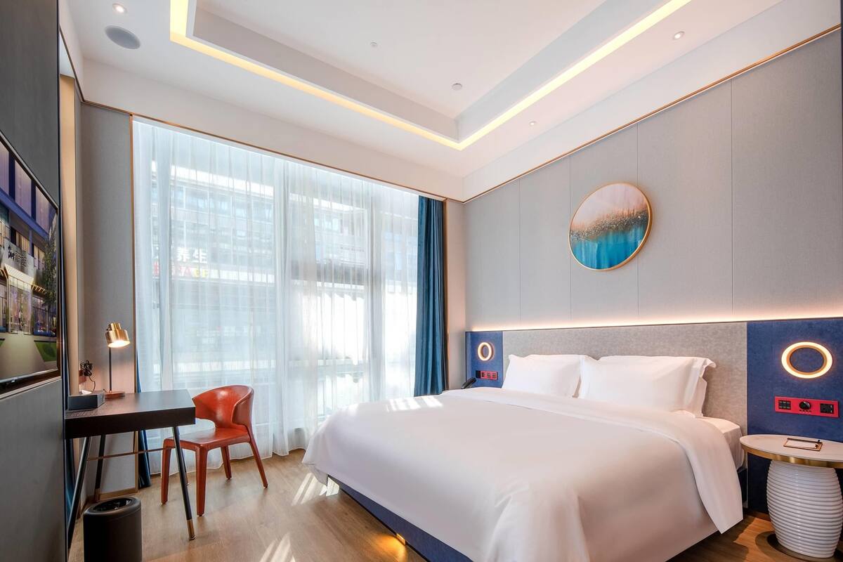 A modern hotel room with a large bed, a desk with a chair, a round table, and a large window with curtains.