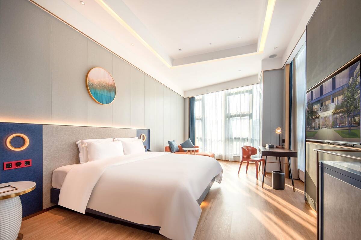 A modern hotel room with a large bed, a desk, a chair, and a view of the city through large windows.