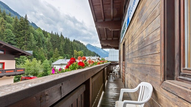 Ein Holzbalkon mit einem weißen Plastikstuhl, der auf eine Berglandschaft mit Häusern und roten Blumen blickt.