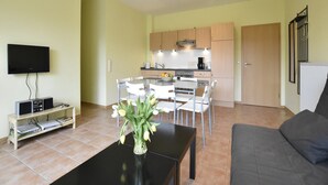 A modern living room with a flat-screen TV, a coffee table with a vase of flowers, a gray sofa, and a kitchen area with wooden cabinets and a dining table. - Küstenwohnung in Zierow (Zierow)
