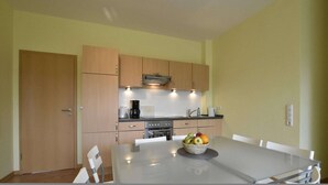 A compact kitchen with wooden cabinets, a stove, and a dining table with a fruit bowl. - Küstenwohnung in Zierow (Zierow)