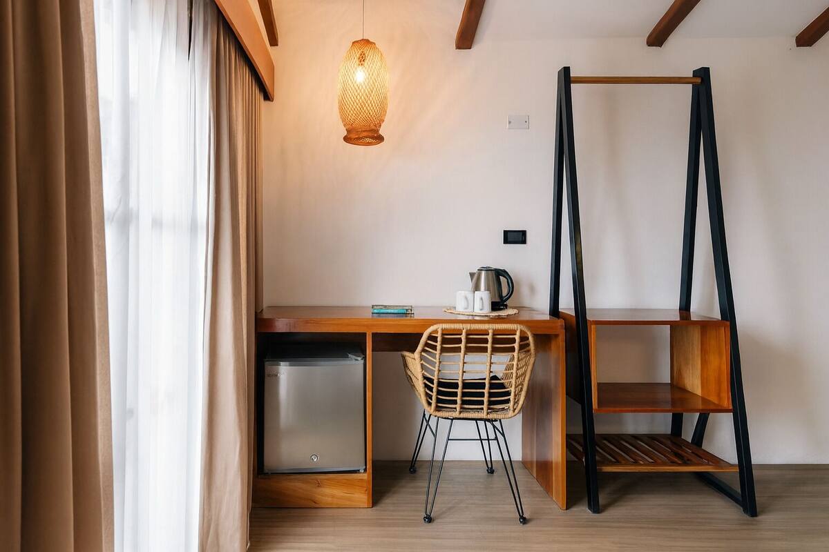 A compact room with a wooden desk, a wicker chair, a hanging light fixture, and a black metal shelf.
