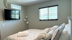 A bedroom with a bed, a flat-screen TV mounted on the wall, a window with blinds, and a ceiling fan.